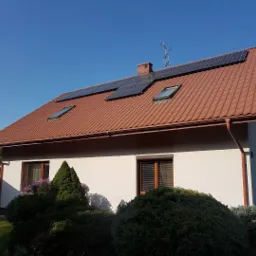 Dom jednorodzinny z brązową dachówką, na której zamontowane są panele słoneczne, widoczne również okna dachowe i antena telewizyjna.