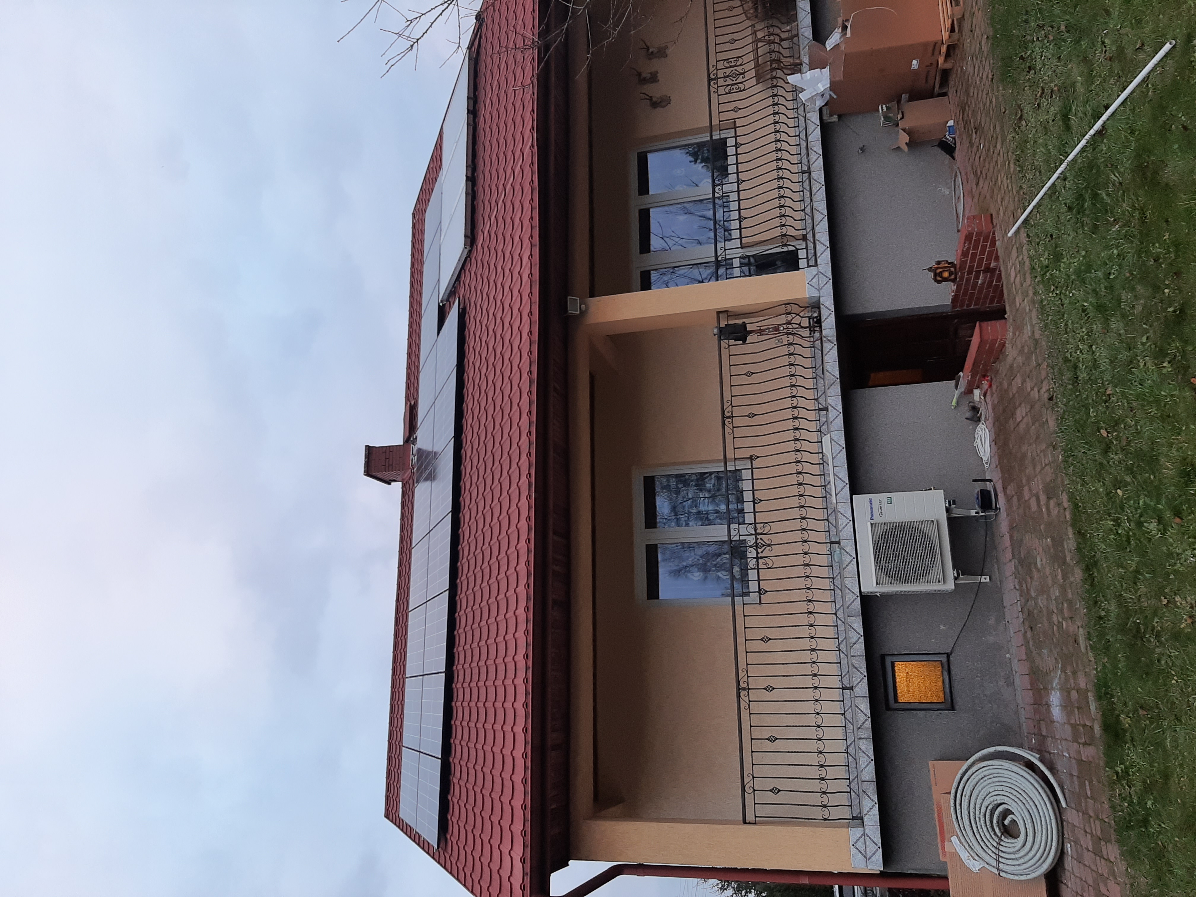 Dom jednorodzinny z panelami fotowoltaicznymi na dachu z czerwonej dachówki, widoczny również klimatyzator na ścianie budynku.