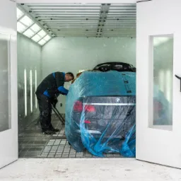 Pracownik w kombinezonie zabezpiecza folią ochronną tylną część czarnego samochodu marki BMW wewnątrz lakierni samochodowej z kratką podłogową.