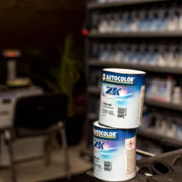 Dwie puszki farby samochodowej Autocolor 2K, jedna na drugiej, w warsztacie lakierniczym, na tle półek z farbami i sprzętu warsztatowego.