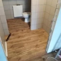 Wykończona podłoga imitująca drewno w małej łazience w trakcie prac, widoczny częściowo zamontowany sedes i płytki na ścianach, brak listew przypodłogowych.