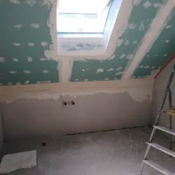 Poddasze ze skośnym sufitem, okno dachowe, ściany pokryte częściowo płytami gipsowo-kartonowymi, widoczne ślady szpachlowania, na podłodze fragment płyty, drabina oparta o ścianę.