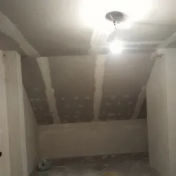 Poddasze w trakcie prac wykończeniowych: widoczne płyty kartonowo-gipsowe na skosach, łączenia wzmocnione taśmą i pokryte masą szpachlową, surowe ściany i sufit, prowizoryczne oświetlenie.