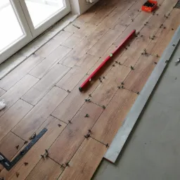 Układanie płytek imitujących drewno na podłodze, widoczne poziomice, krzyżyki dystansowe i pudełko po krzyżykach, częściowo ułożona podłoga w pomieszczeniu z oknem.
