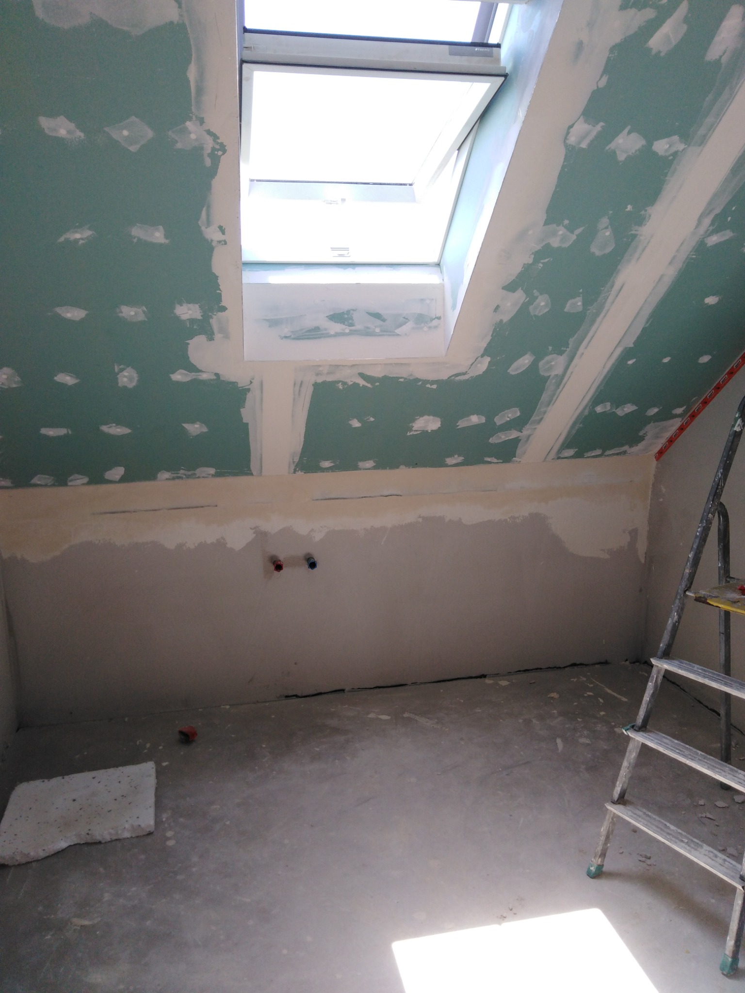 Poddasze ze skośnym sufitem, okno dachowe, ściany pokryte częściowo płytami gipsowo-kartonowymi, widoczne ślady szpachlowania, na podłodze fragment płyty, drabina oparta o ścianę.