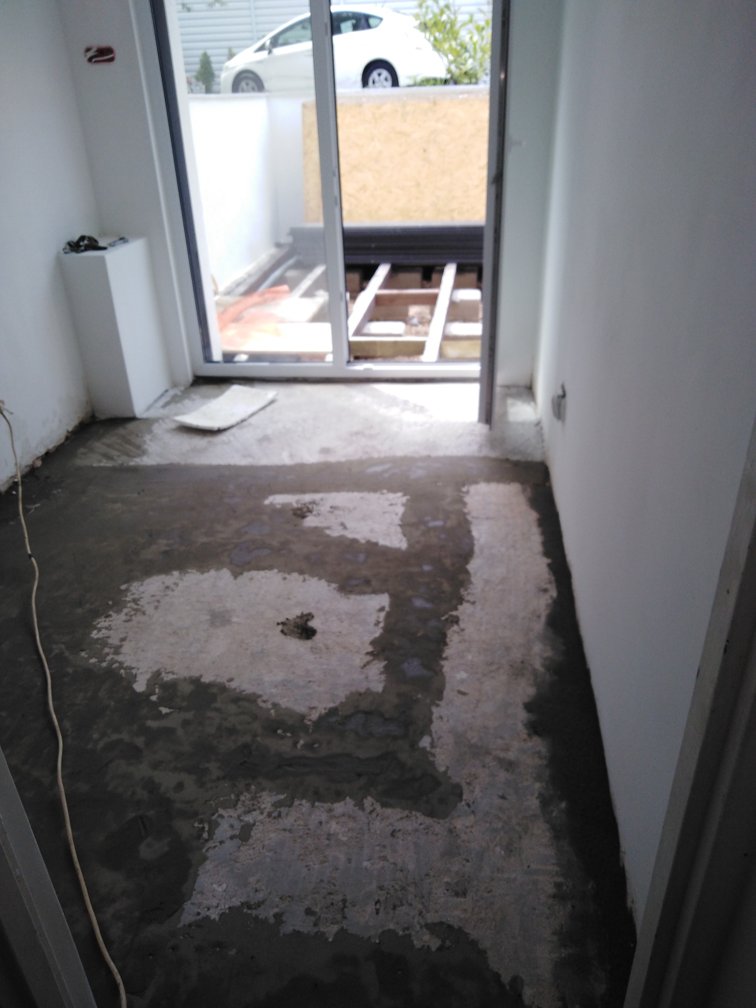 Wylewka podłogowa w trakcie schnięcia w pomieszczeniu z dużym oknem przesuwnym, widoczne ślady wilgoci i nierówności na powierzchni betonu, w tle konstrukcja podłogi i fragment białego samochodu za...