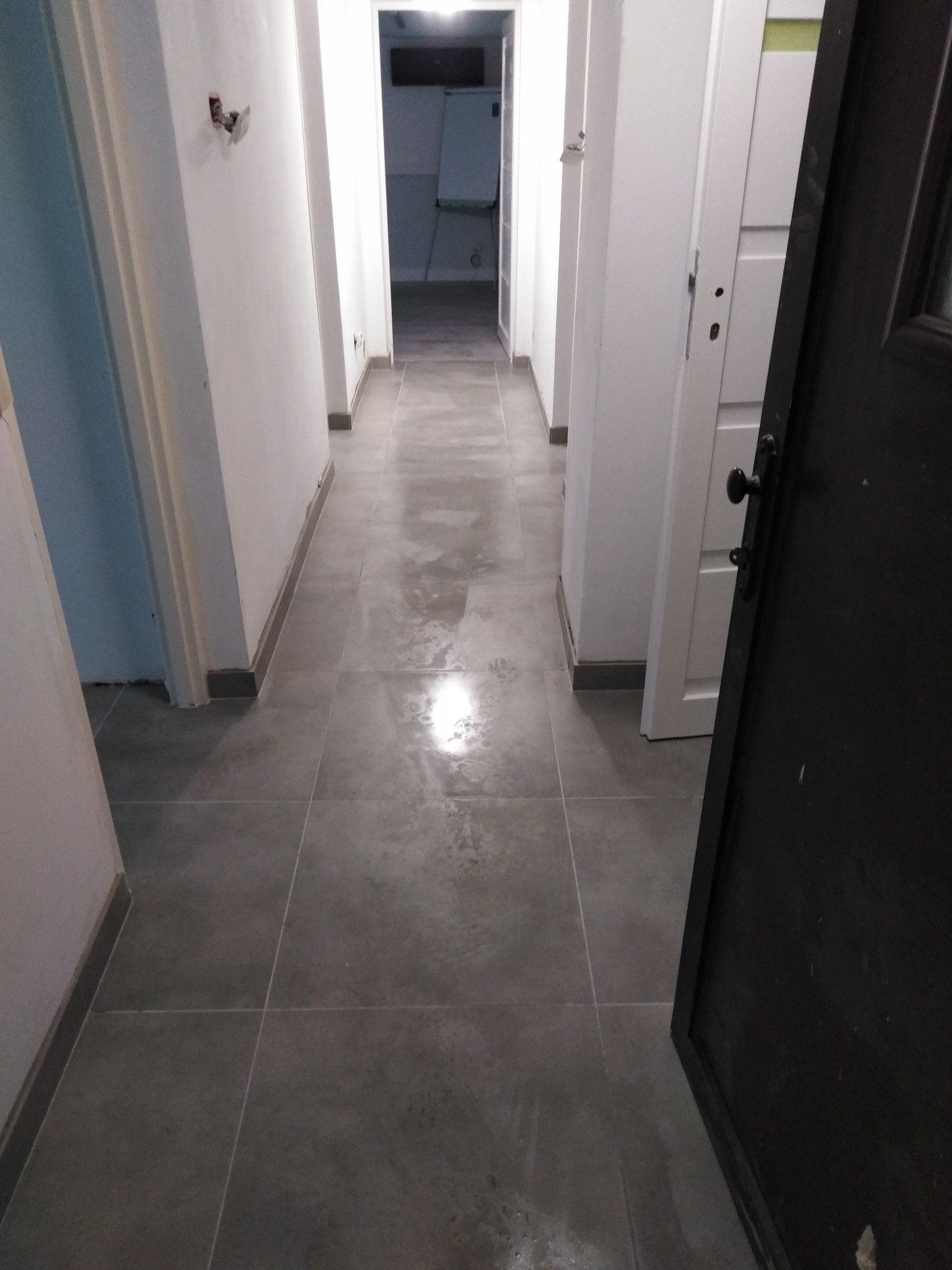 Wyremontowany korytarz z szarymi płytkami podłogowymi, białe ściany i framugi drzwiowe, widoczne ślady po niedawnych pracach, w tle widoczna kuchnia.