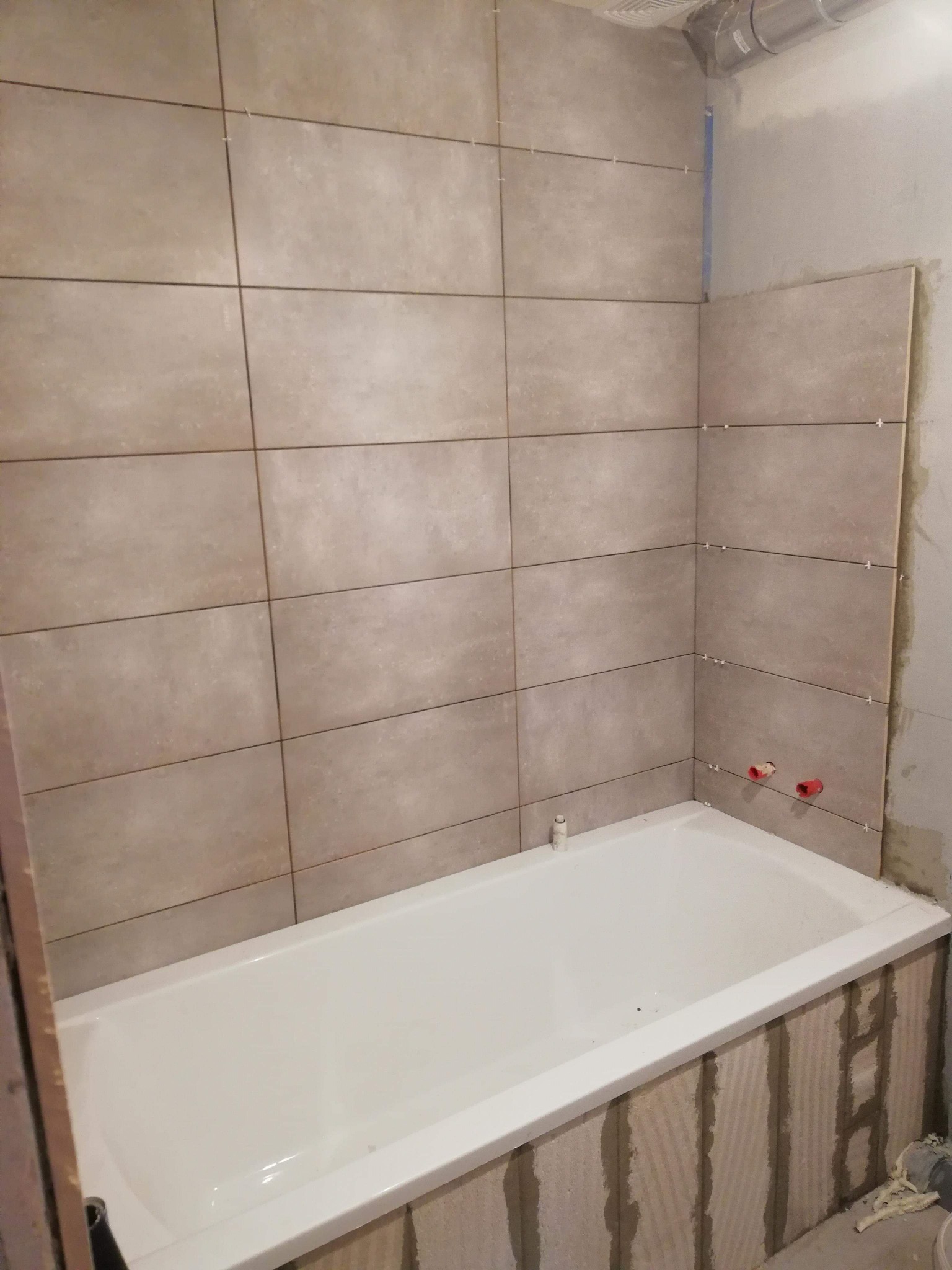 Jasne, prostokątne płytki ceramiczne ułożone na ścianie nad białą wanną w trakcie remontu łazienki, widoczne krzyżyki dystansowe między płytkami i niezakończone obłożenie wanny płytkami.