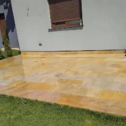 Nowoczesny taras wyłożony polerowanymi płytami w odcieniach żółci i beżu, przylegający do jasnoszarego budynku z brązowymi oknami i drzwiami, z widocznym fragmentem trawnika i tujami w tle.