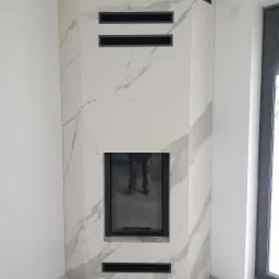 Nowoczesny kominek narożny wykończony płytami imitującymi marmur, z czarnymi kratkami wentylacyjnymi i wkładem kominkowym z widocznym odbiciem nóg fotografa.