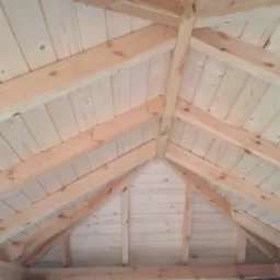 Wnętrze drewnianej konstrukcji z widocznym dachem z desek i belek, surowe drewno bez wykończenia, widok z dołu ukazujący geometrię konstrukcji.