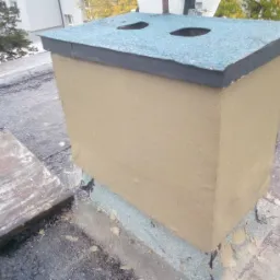 Otynkowana podstawa komina z dwoma otworami wentylacyjnymi na płaskim dachu pokrytym papą.