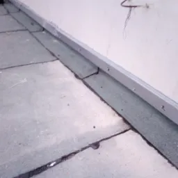 Fragment dachu pokrytego papą z metalową listwą przy ścianie, widoczne łączenia papy zalane masą bitumiczną.