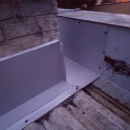 Szare, metalowe elementy wykończeniowe dachu, zamocowane śrubami, widoczne na tle starej, zniszczonej powierzchni dachu i fragmentu ściany budynku.