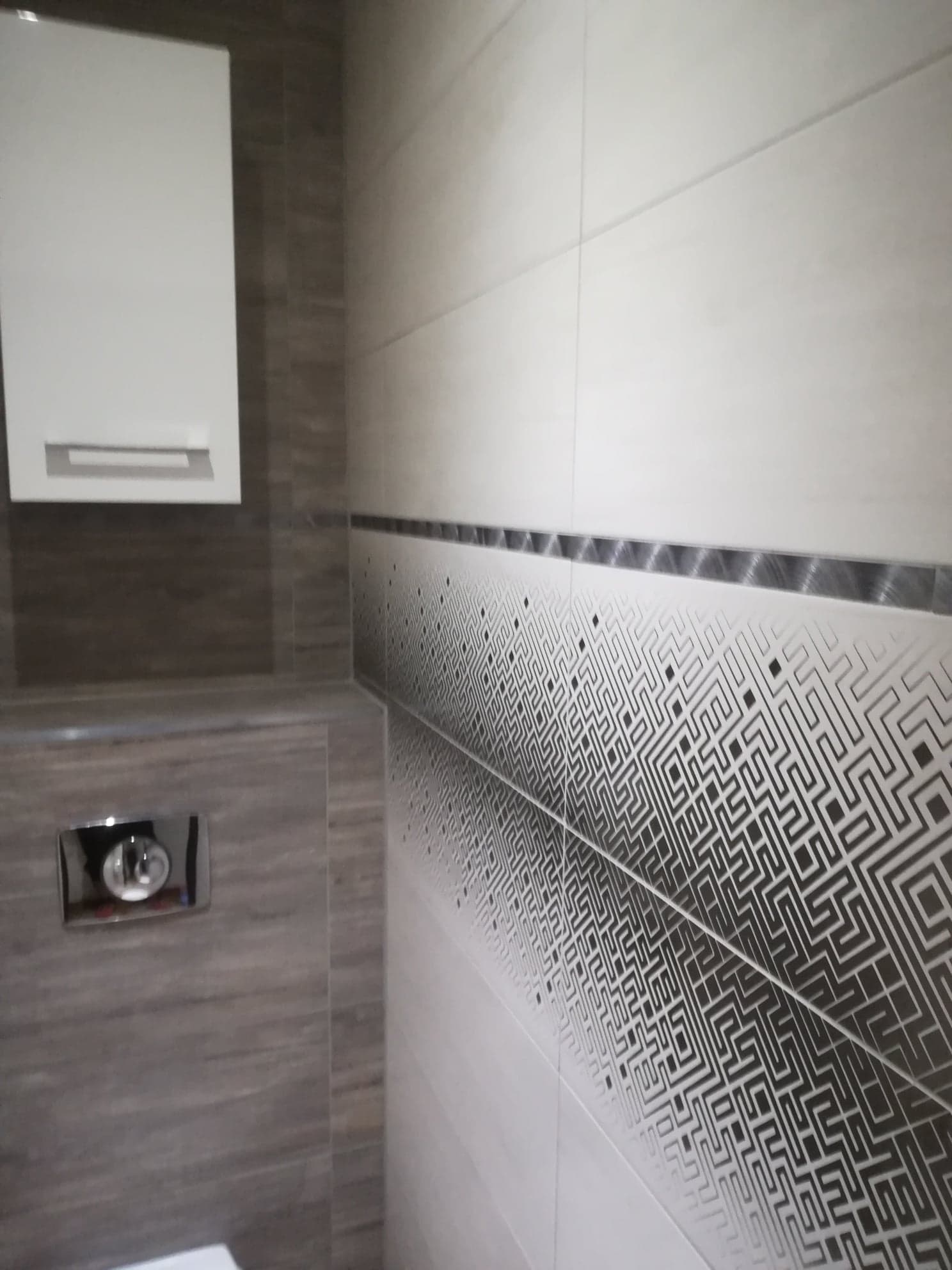 Wykończona łazienka z brązowo-białymi płytkami o wzorze labiryntu, zabudowana toaleta z chromowanym przyciskiem spłukiwania i wisząca szafka.