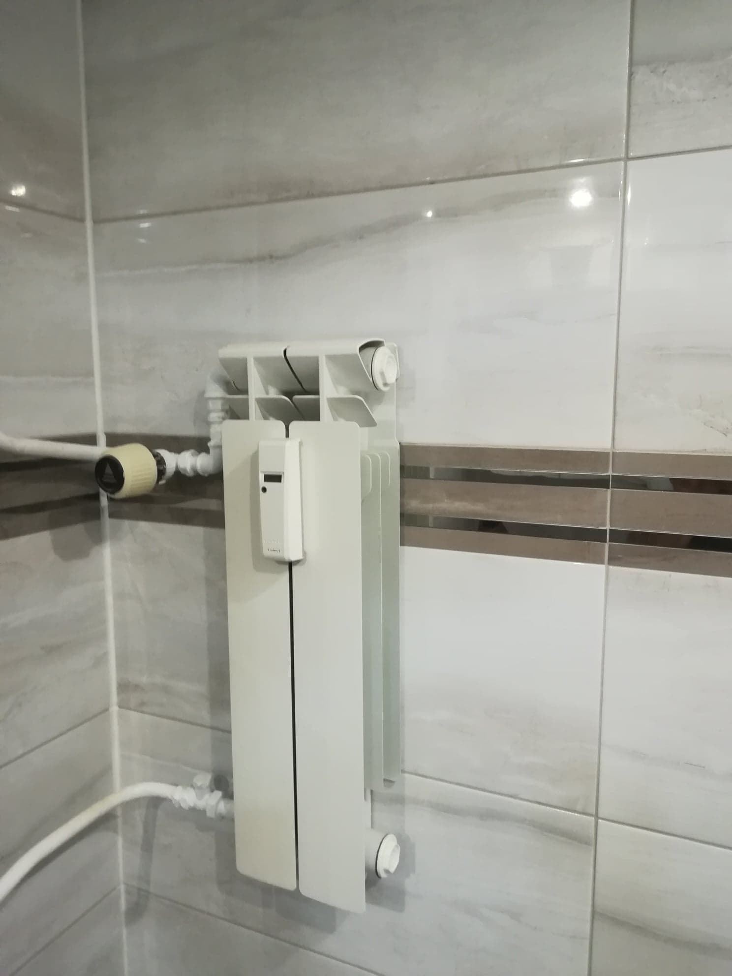 Biały grzejnik panelowy z termostatem elektronicznym zamontowany na ścianie wyłożonej płytkami w odcieniach szarości i bieli.