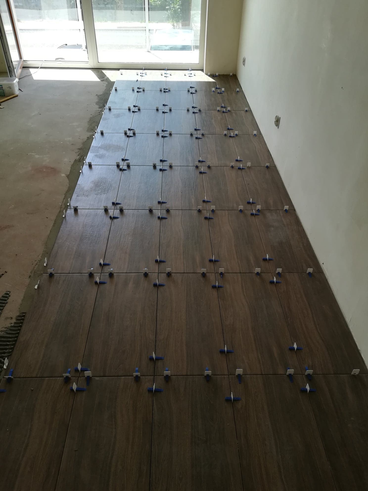 Układanie brązowych płytek podłogowych wewnątrz pomieszczenia, widoczne niebieskie krzyżyki dystansowe, surowy stan podłoża przy oknie.