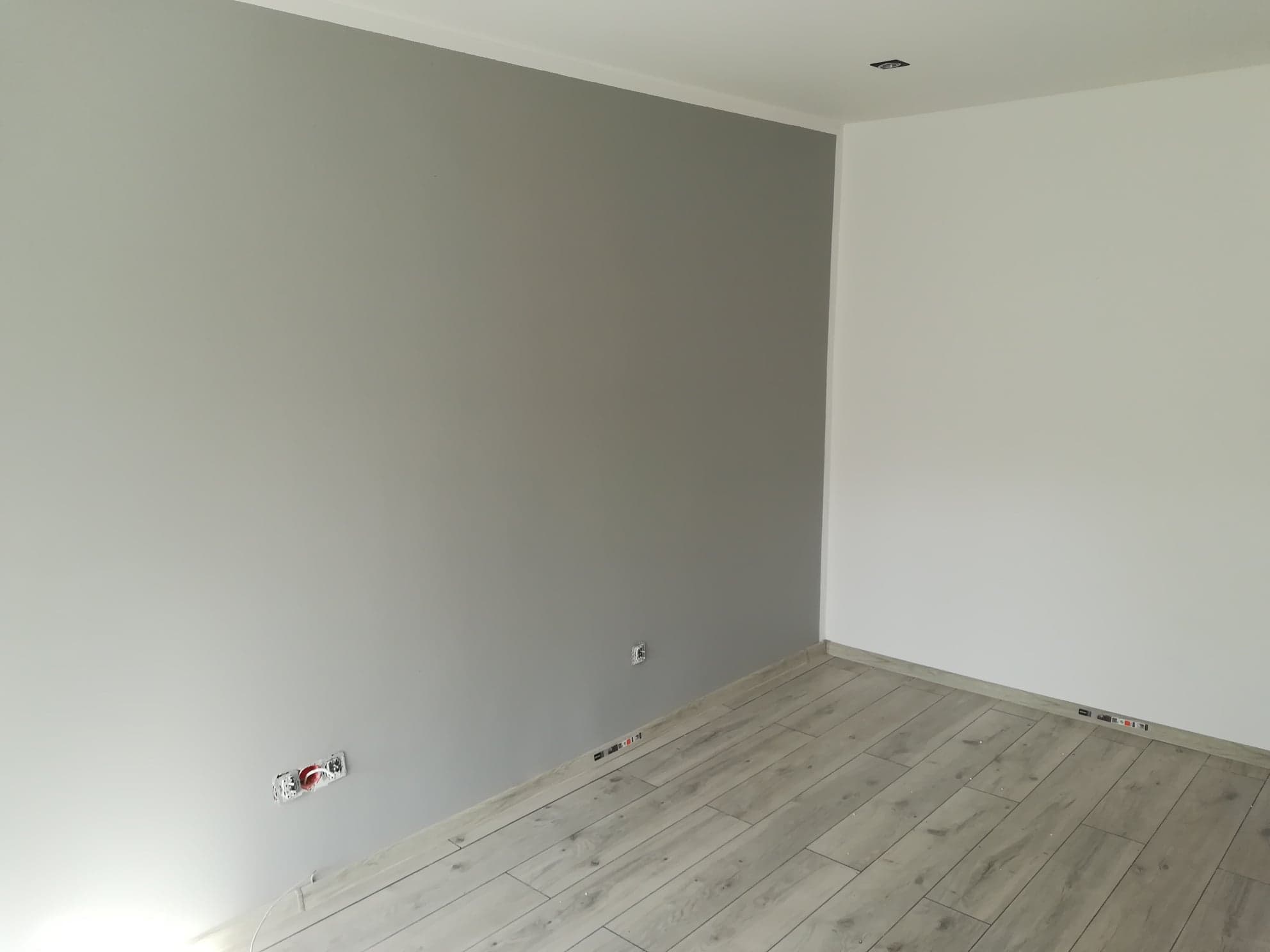 Pokój po remoncie: ściana pomalowana na szaro kontrastuje z białą ścianą, widoczne gniazdka elektryczne i podłoga wyłożona panelami imitującymi drewno w odcieniach szarości.