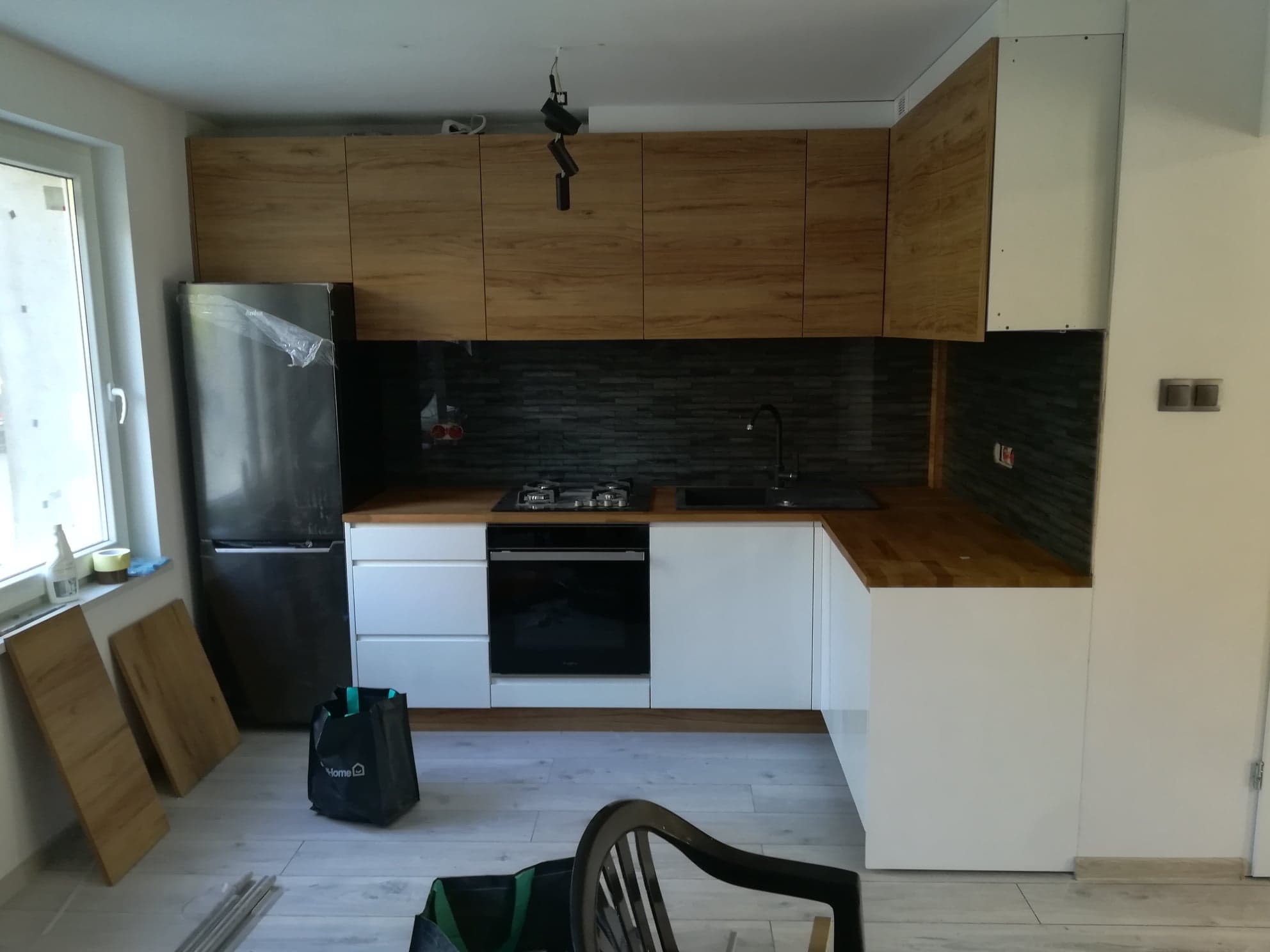 Montaż nowoczesnej kuchni w Gołuskach: białe szafki dolne z drewnianym blatem, drewniane szafki górne, czarny backsplash i lodówka w folii ochronnej przed pierwszym użyciem.