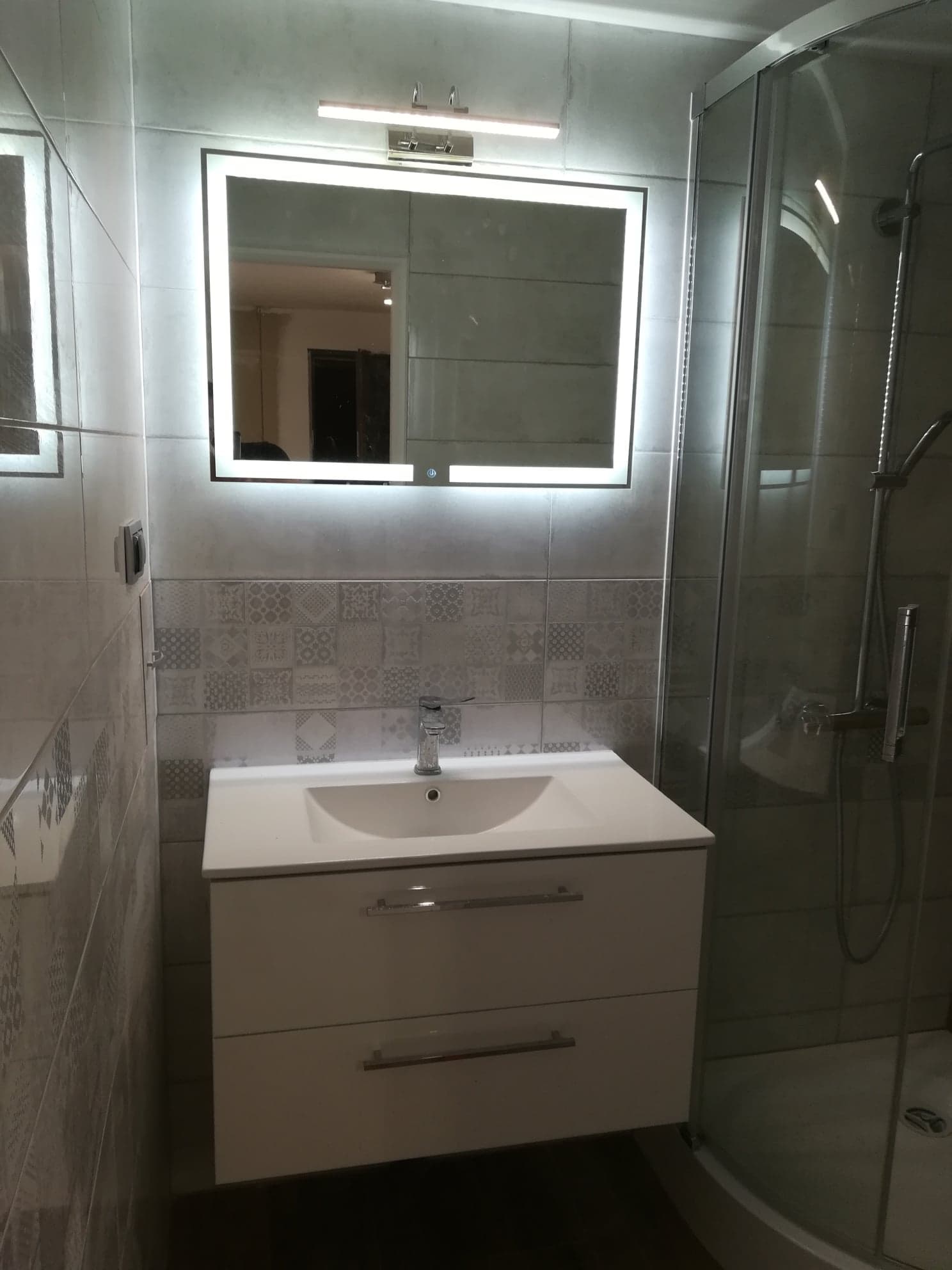 Nowoczesna łazienka z białą szafką pod umywalkę, podświetlanym lustrem i kabiną prysznicową ze szklanymi drzwiami. Ściany wyłożone szarą płytką z dekoracyjnym pasem. Widoczny fragment baterii...