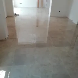 Wyłożona jasnobeżowymi płytkami podłoga w trakcie remontu, widoczne niedokończone ściany i wystające przewody elektryczne.