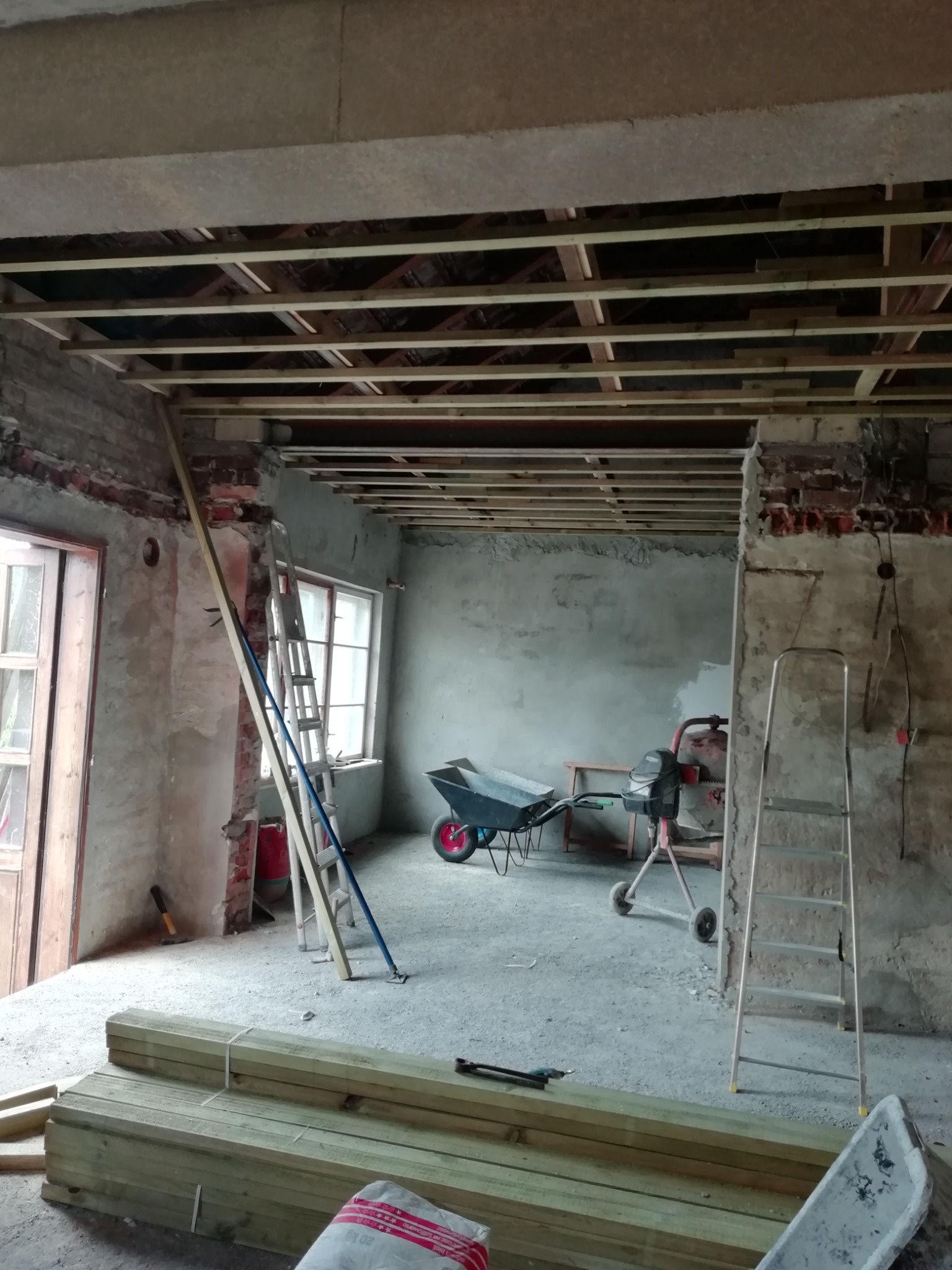 Wnętrze w trakcie remontu: odsłonięta konstrukcja sufitu z drewnianych belek, surowy tynk, drabiny, taczka i betoniarka w pomieszczeniu z przygotowanymi deskami.