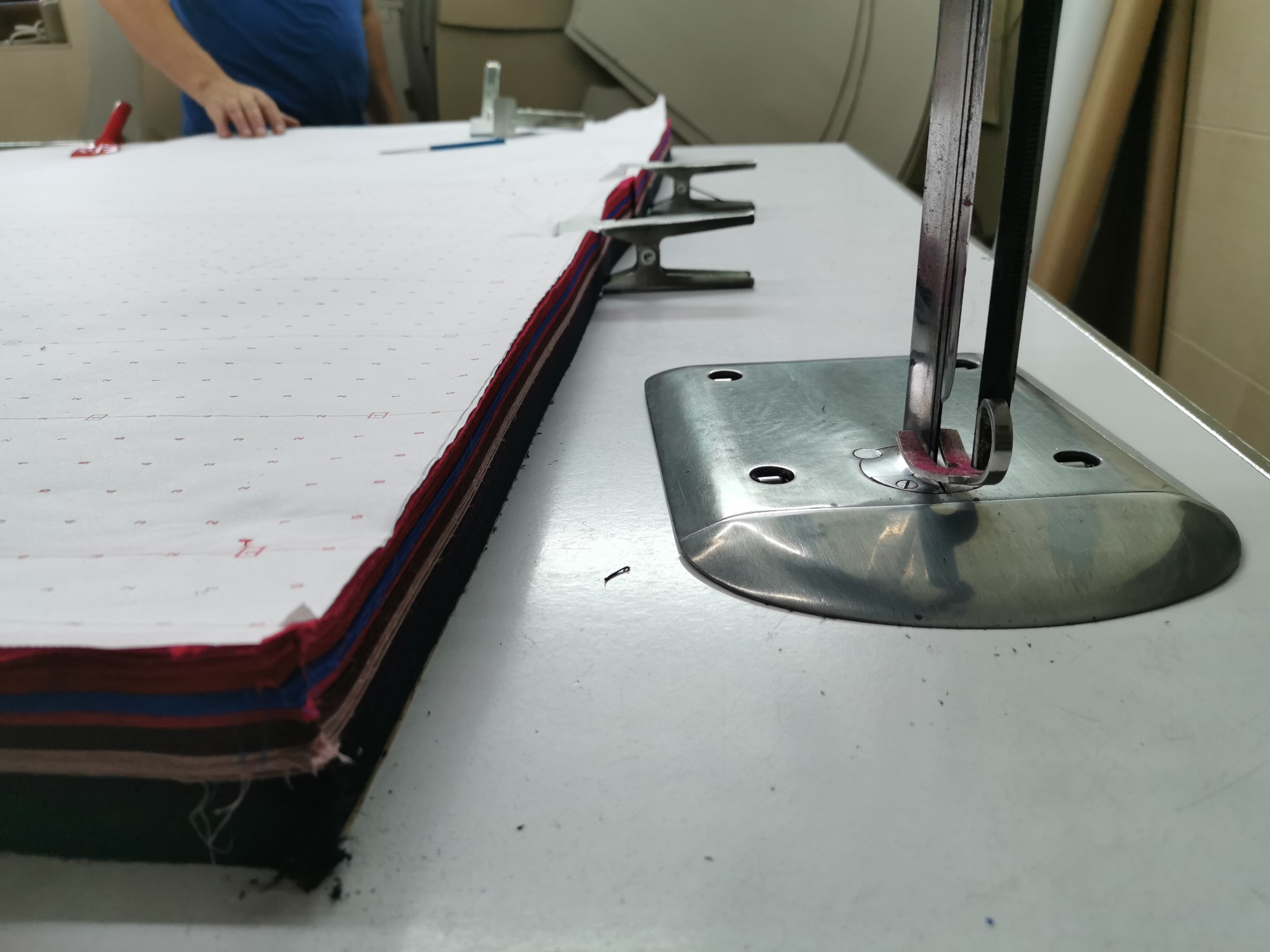 Wielowarstwowa sterta tkanin z naniesionym wzorem wykroju, zabezpieczona klipsami, przy krawędzi stołu z maszyną do cięcia.