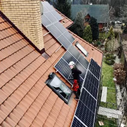 Montera fotowoltaiki w kasku i uprzęży na dachu z dachówki ceramicznej, instalujący panele słoneczne obok okna dachowego; widoczne rusztowanie i komin z cegły klinkierowej.