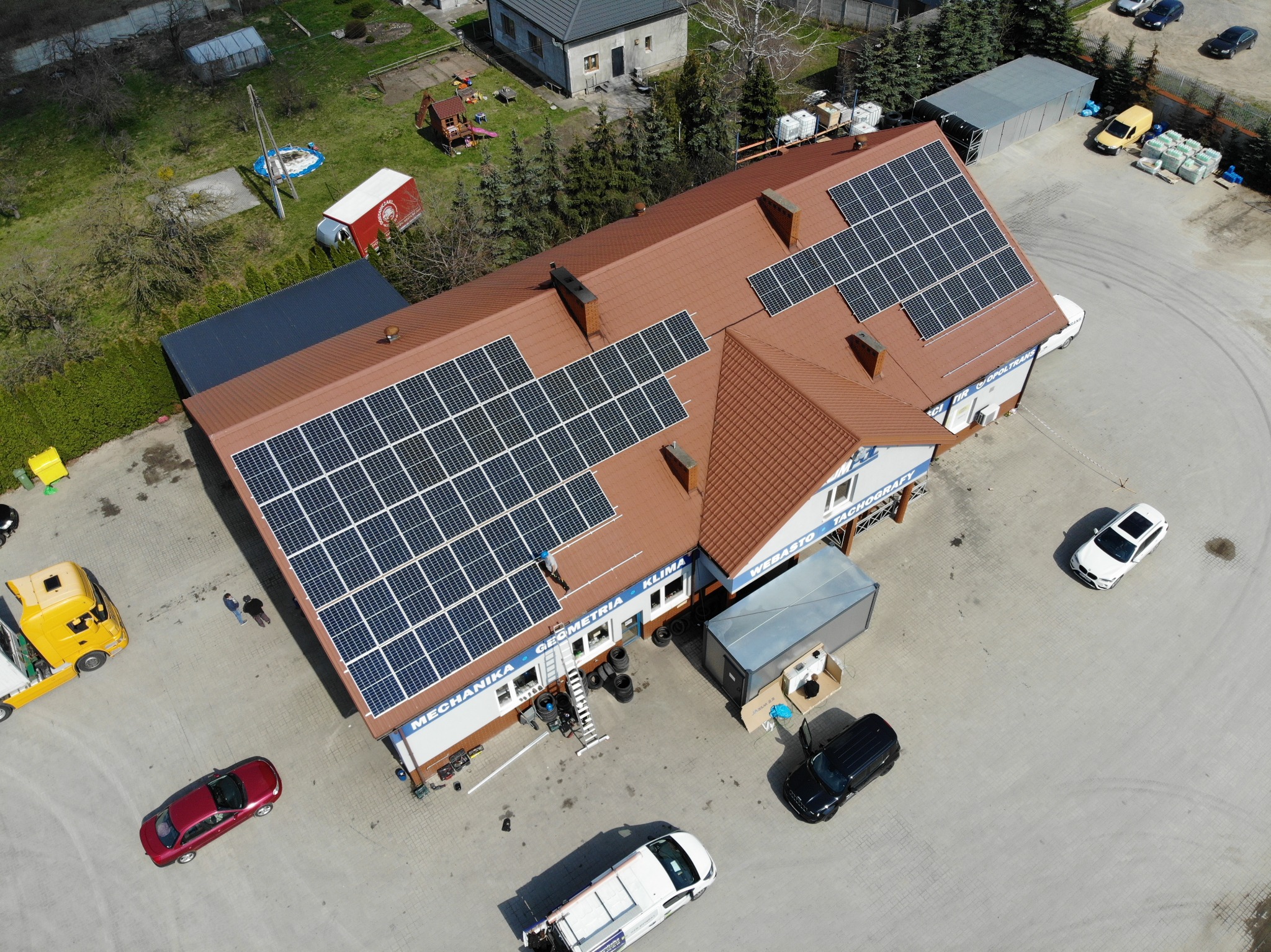 Widok z drona na budynek warsztatu samochodowego z panelami słonecznymi na dachu, zaparkowanymi samochodami i żółtą lawetą.