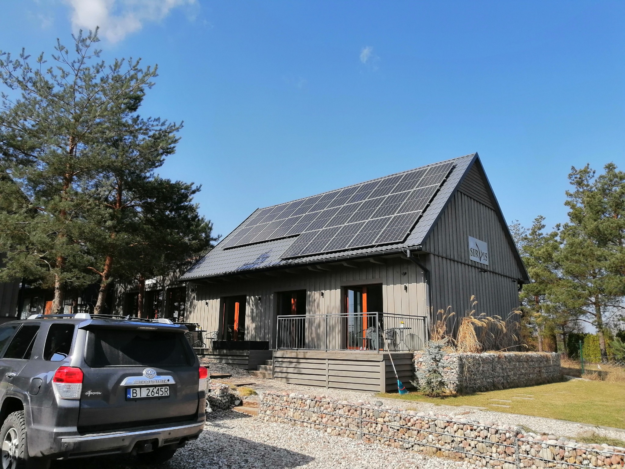 Nowoczesny dom z szarą elewacją i panelami słonecznymi na dachu, widok z zewnątrz na jasny dzień, z zaparkowanym szarym SUV-em na żwirowym podjeździe.