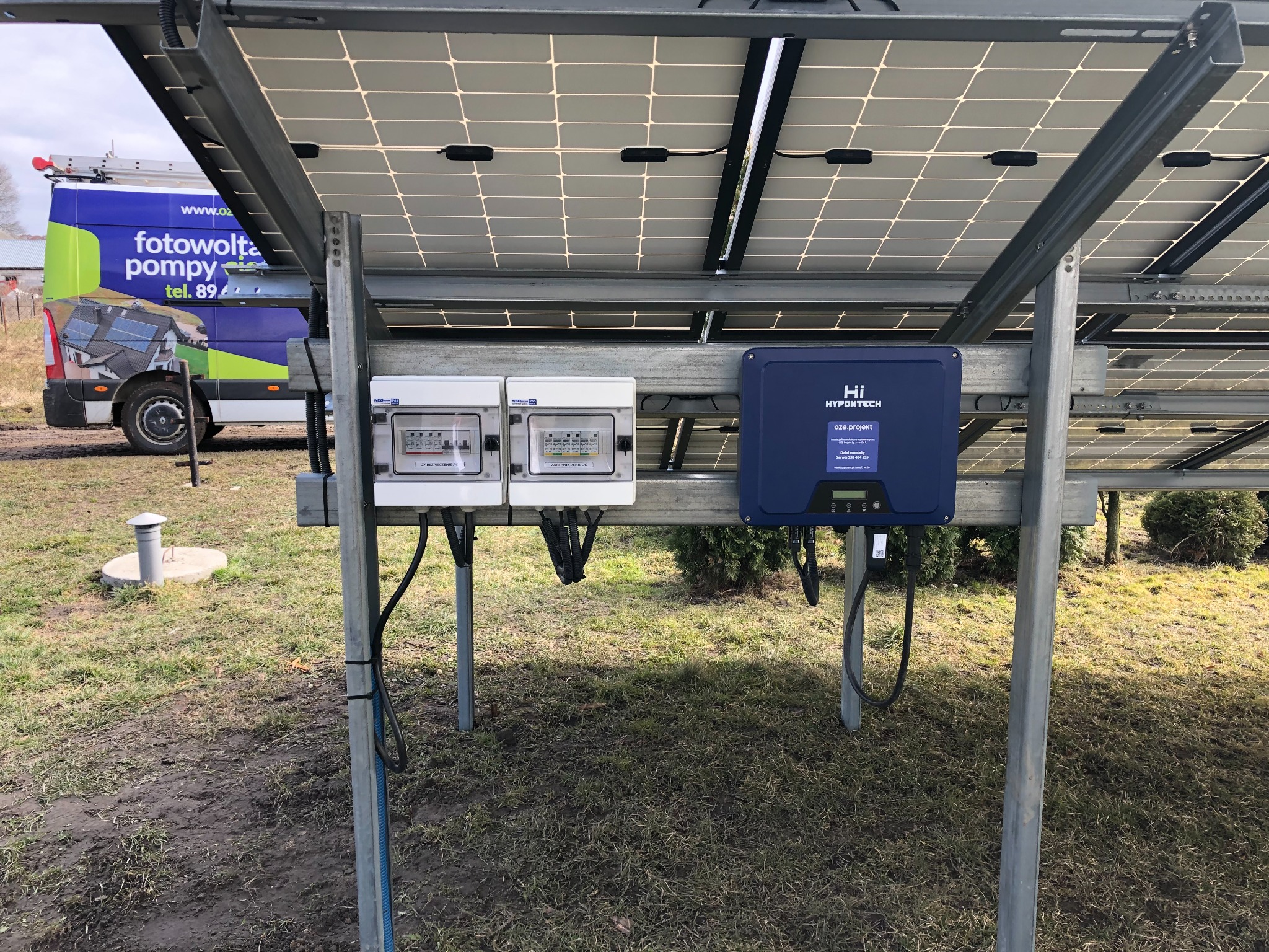 Instalacja paneli słonecznych z widocznymi dwoma skrzynkami zabezpieczeń i falownikiem Hypontech na metalowej konstrukcji, w tle samochód dostawczy z reklamą firmy.