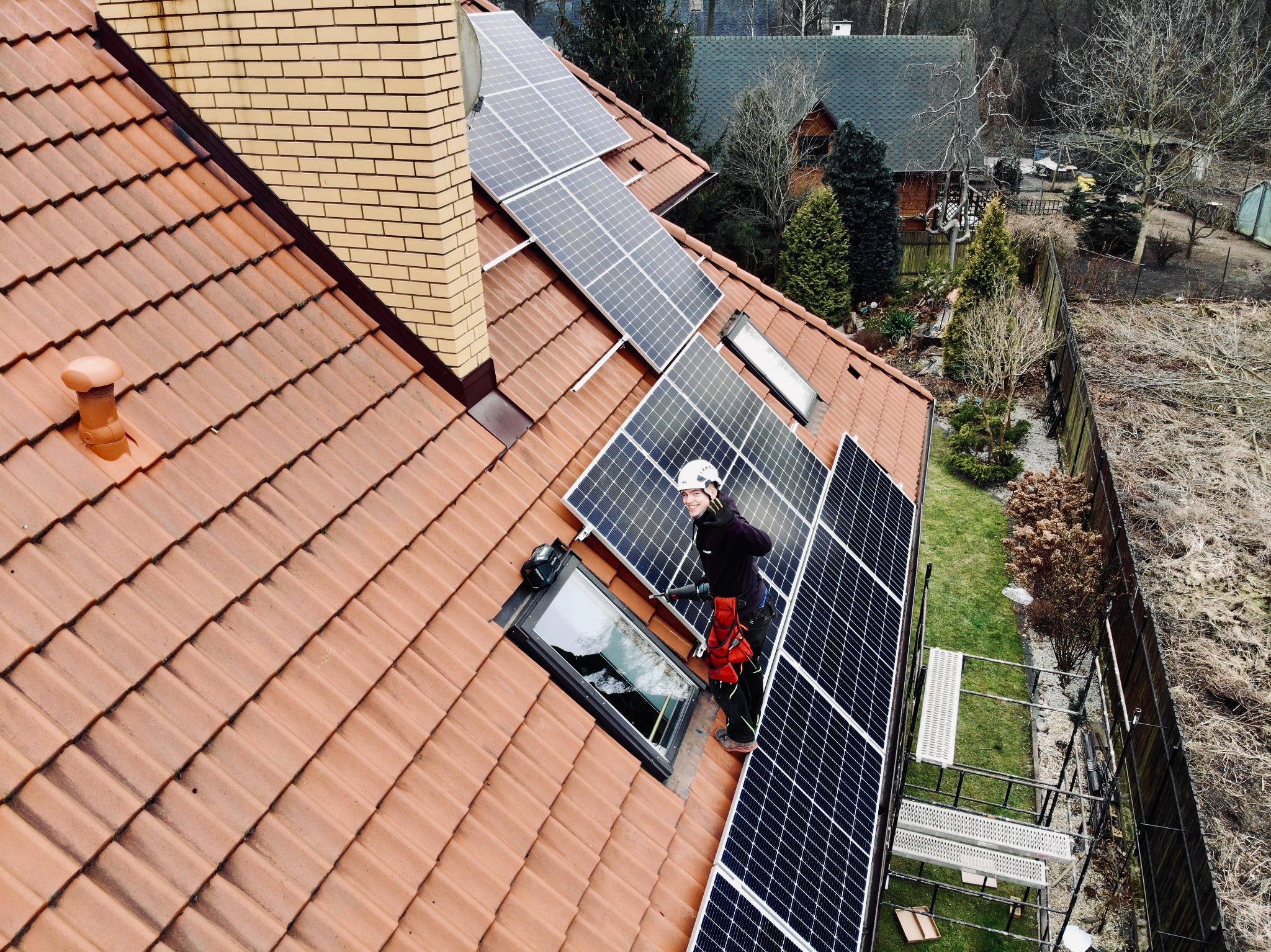 Montera fotowoltaiki w kasku i uprzęży na dachu z dachówki ceramicznej, instalujący panele słoneczne obok okna dachowego; widoczne rusztowanie i komin z cegły klinkierowej.