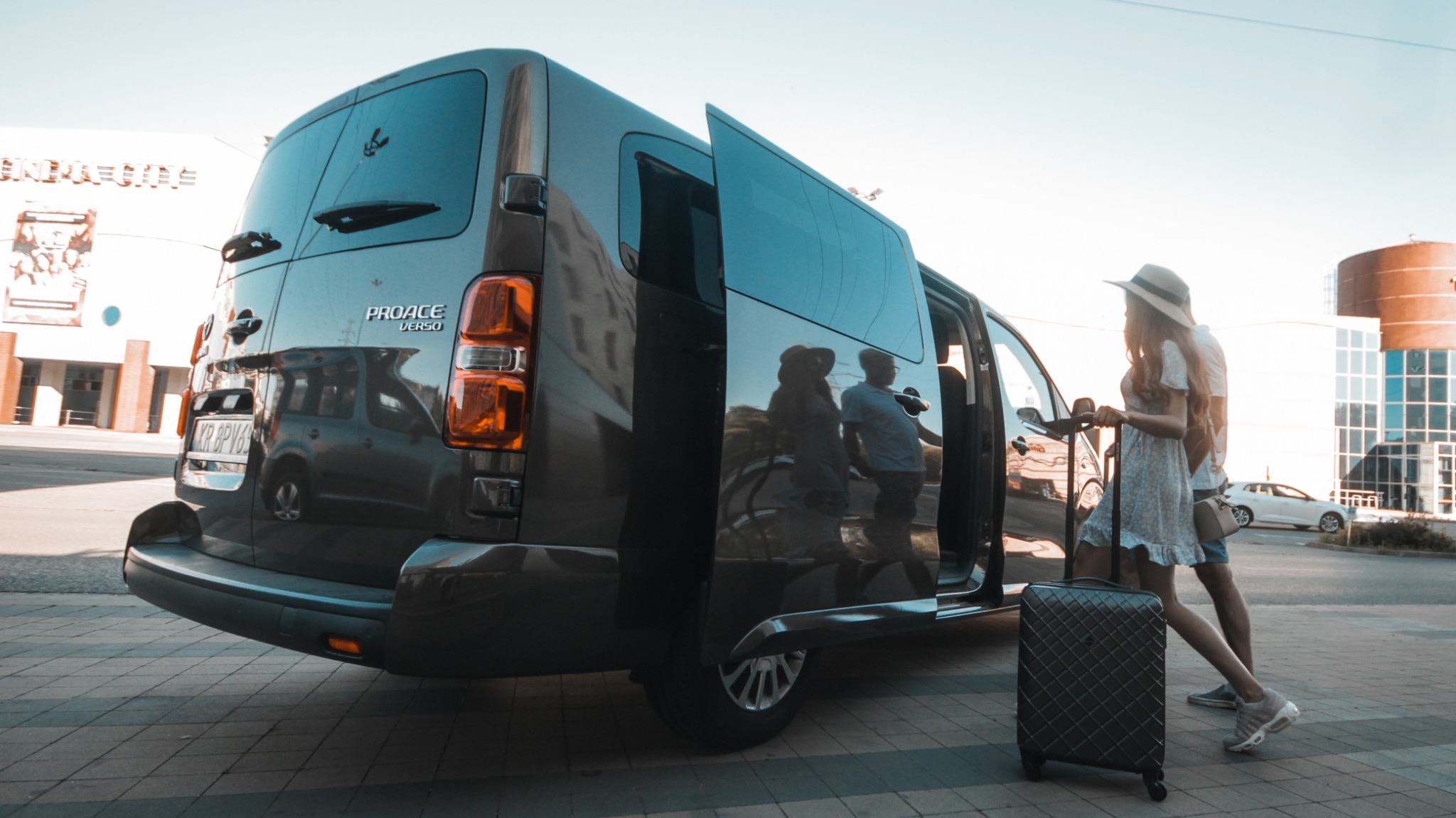 Para z walizką wsiada do ciemnoszarego vana Peugeot Proace Verso z otwartymi drzwiami przesuwnymi, na tle budynku kina.