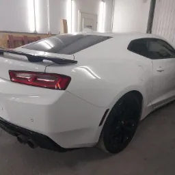 Biały Chevrolet Camaro po lakierowaniu, widok z tyłu, czarne felgi i spojler, wnętrze warsztatu z widoczną ścianą i drzwiami.