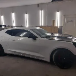 Biały sportowy samochód w trakcie naprawy w warsztacie, zdemontowany przedni zderzak, czarna maska, widoczne oświetlenie warsztatowe