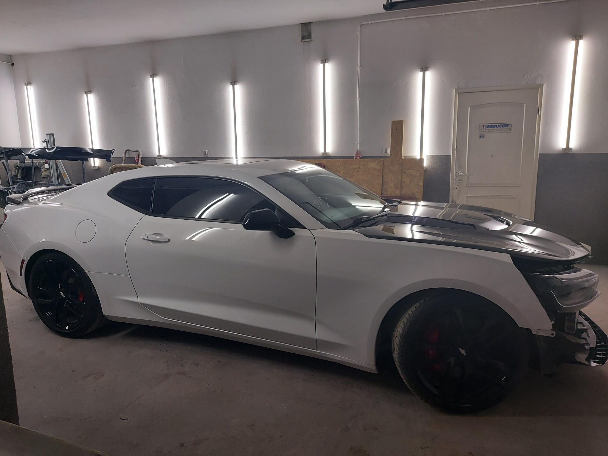 Biały sportowy samochód w trakcie naprawy w warsztacie, zdemontowany przedni zderzak, czarna maska, widoczne oświetlenie warsztatowe