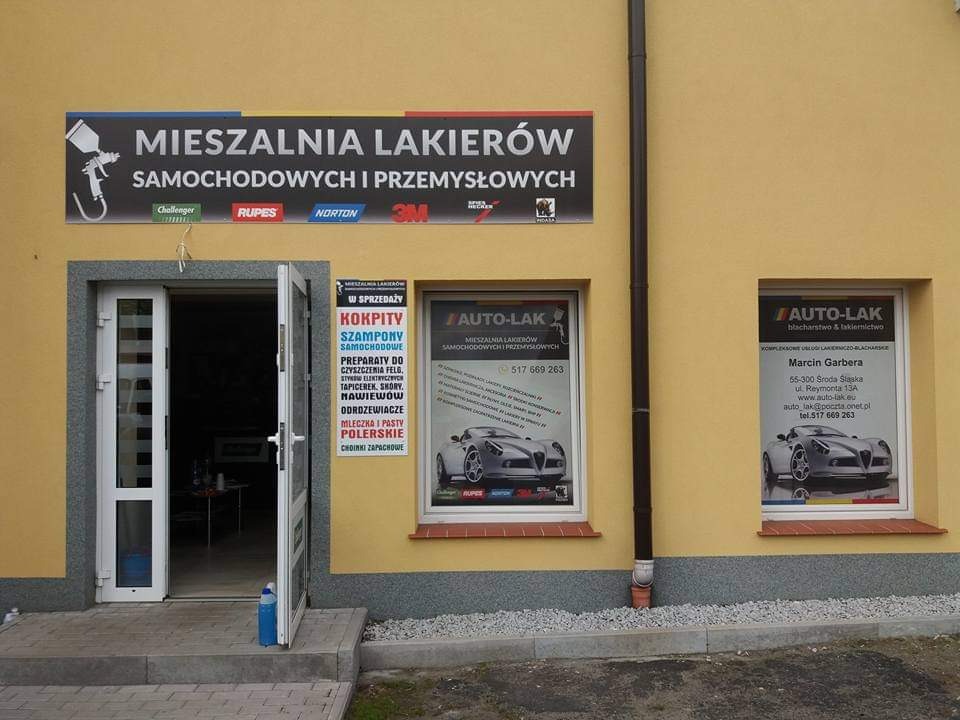 Fasada budynku mieszalni lakierów samochodowych i przemysłowych Auto-Lak z otwartymi drzwiami wejściowymi i oknami z reklamami, widoczny szyld z nazwą i logotypami producentów materiałów...