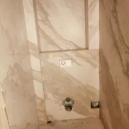 Ściany łazienki wyłożone płytkami imitującymi marmur, widoczne gniazdko elektryczne i rura kanalizacyjna przed montażem armatury, żółte taśmy zabezpieczające.