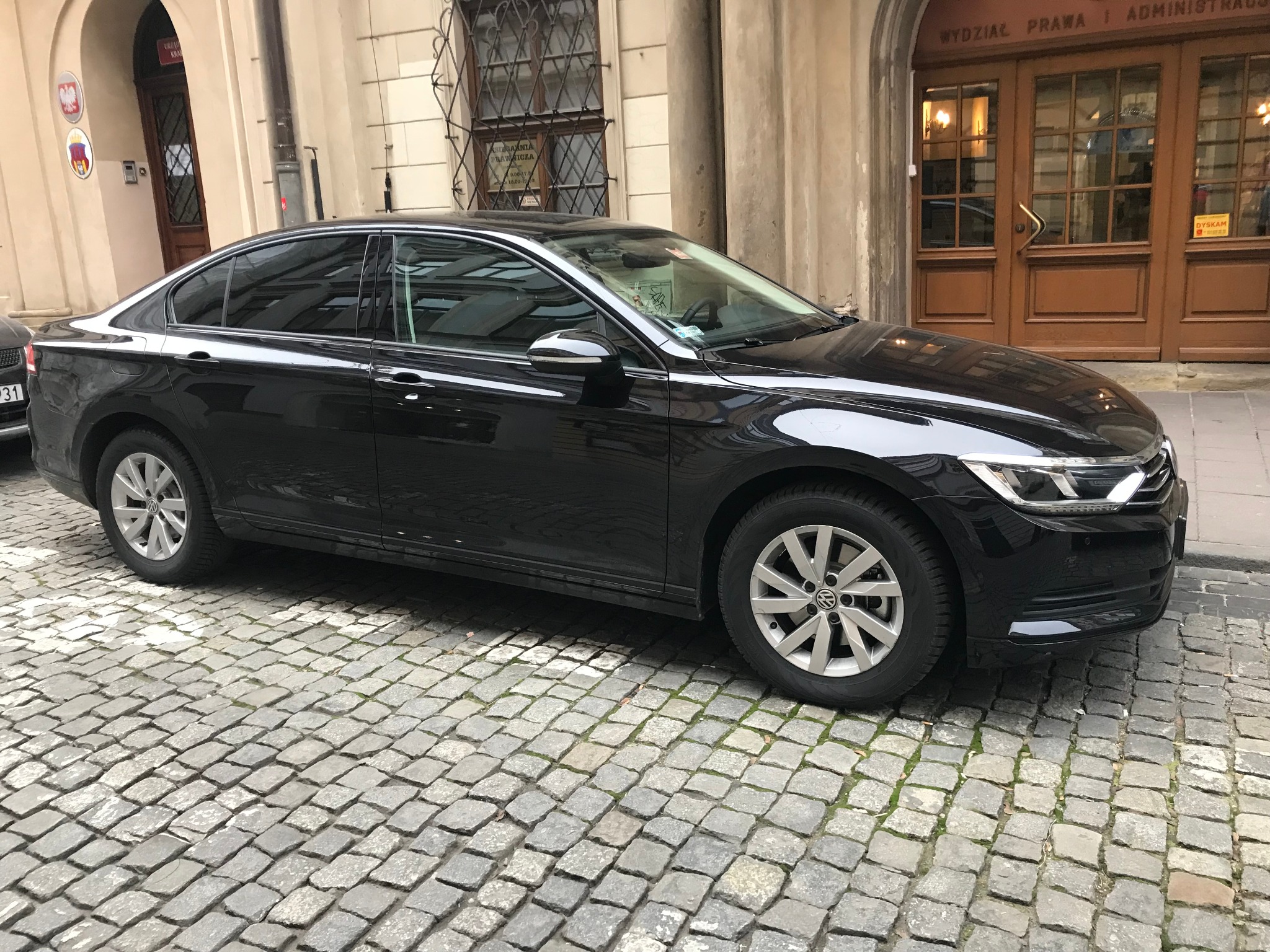 Czarny VW Passat zaparkowany na brukowanej ulicy przed budynkiem Wydziału Prawa i Administracji, widok z boku.