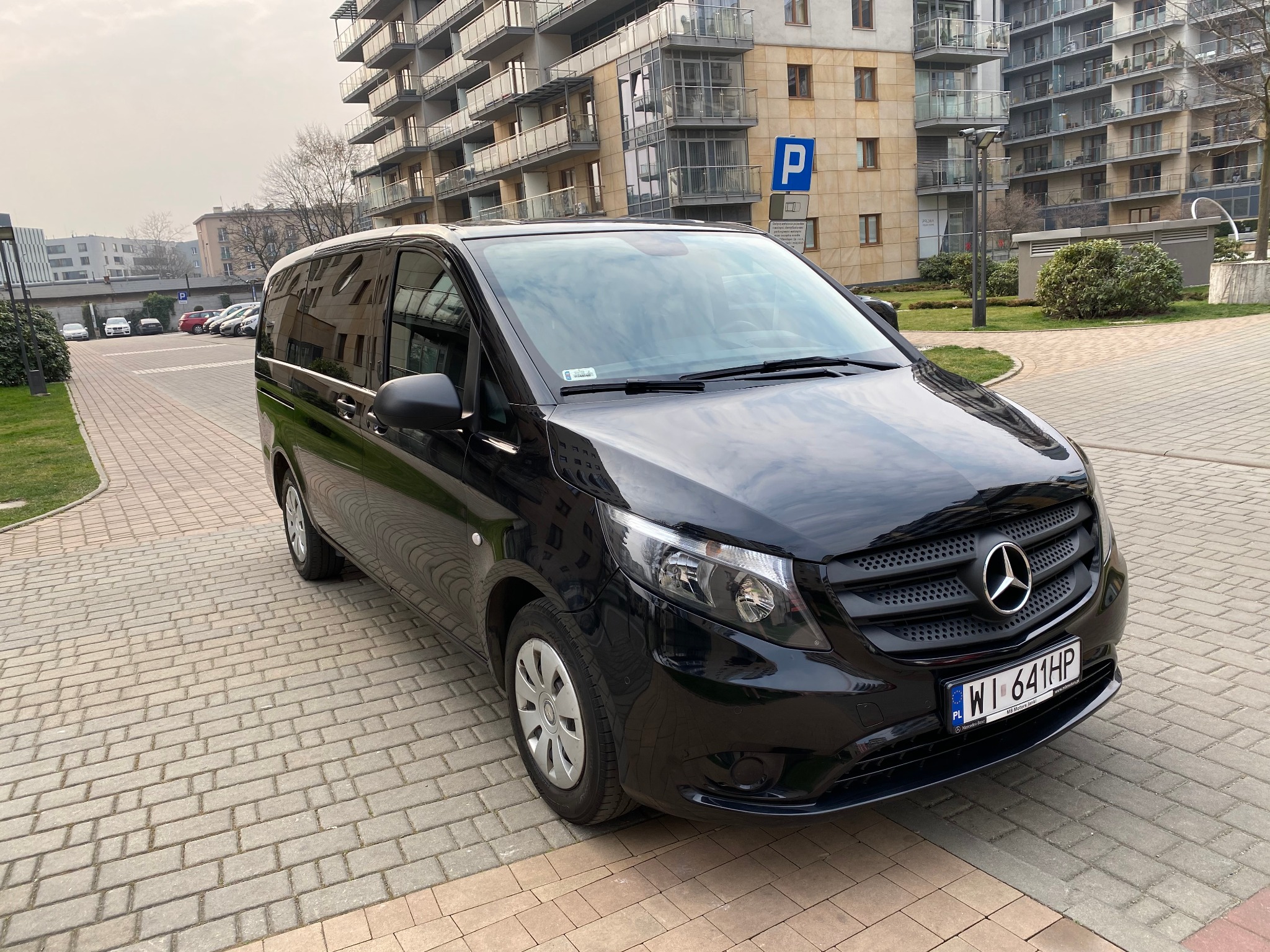 Czarny Mercedes Vito zaparkowany na brukowanej nawierzchni przed nowoczesnym budynkiem mieszkalnym z balkonami; widoczny znak parkingu.