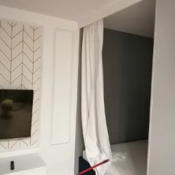 Pokój po remoncie: ściana z tapetą w geometryczny wzór, biała zabudowa meblowa, zasłonięte łóżko za białą kotarą, widoczna mop w wiadrze.