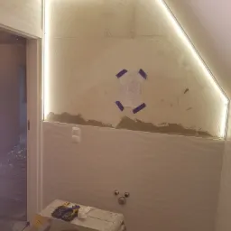 Łazienka w trakcie remontu: przygotowana ściana pod glazurę z dekoracyjnym oświetleniem LED, widoczne narzędzia na drabince malarskiej, instalacja wodna i schemat techniczny przyklejony do ściany.