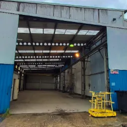 Szeroka brama wjazdowa do hali przemysłowej z szarej blachy falistej z niebieskim dołem, widoczne wnętrze hali z suwnicą, na zewnątrz stoją kontenery i podnośnik.