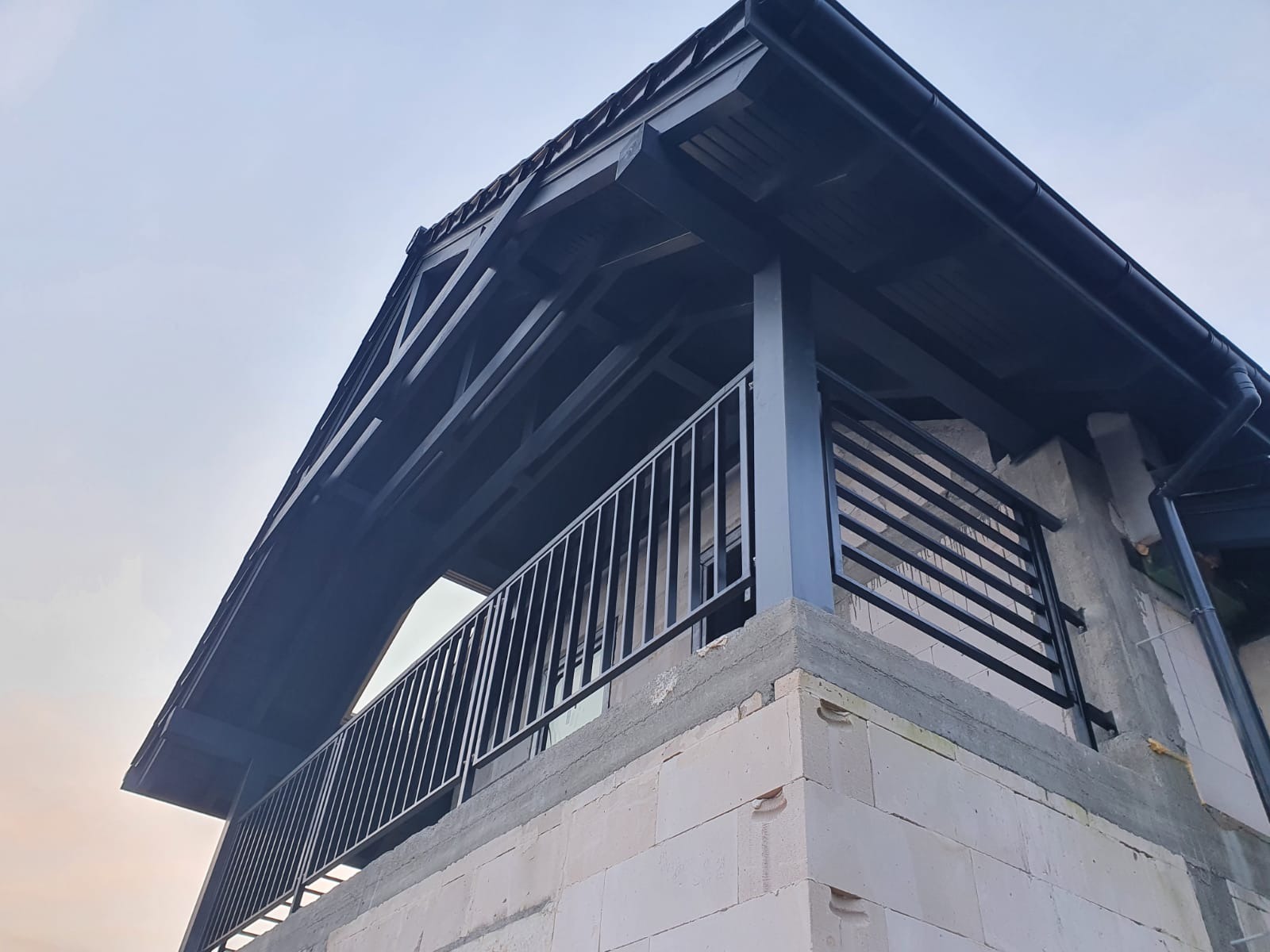 Nisko kątowe ujęcie balkonu z czarną metalową balustradą o pionowych szczeblach, wspartego na murze z białych bloczków, z widocznym fragmentem dachu i niebem w tle.