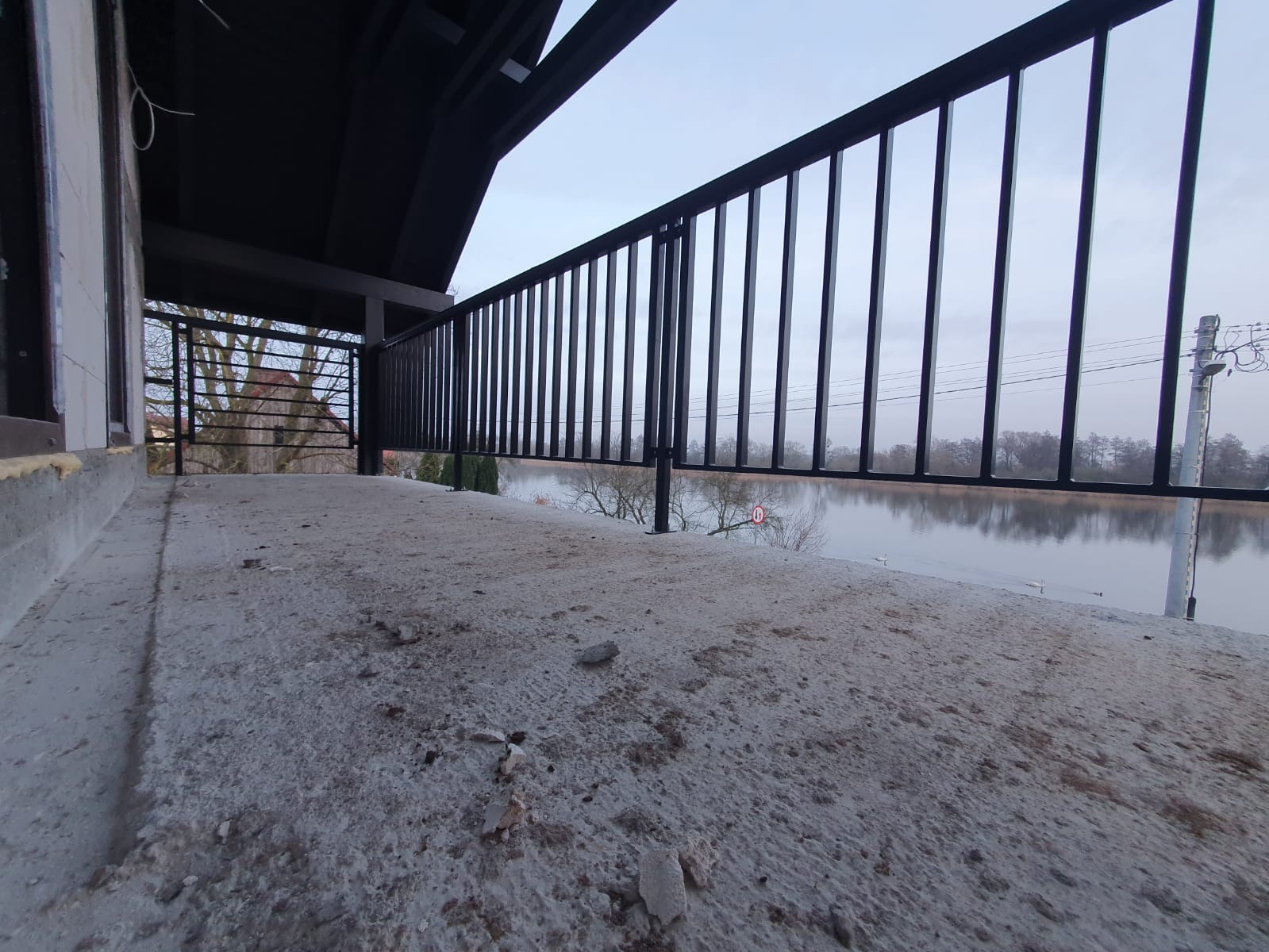 Czarna, metalowa balustrada na betonowym balkonie w trakcie budowy, z widokiem na jezioro i budynek w tle.