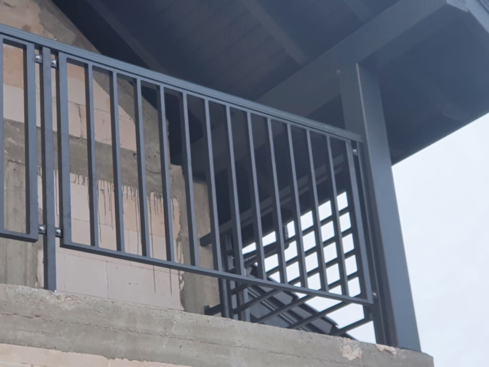 Szara, stalowa balustrada balkonowa z pionowymi prętami i kratką w dolnej części, zamontowana na surowym, betonowym balkonie w trakcie budowy, widok z dołu.