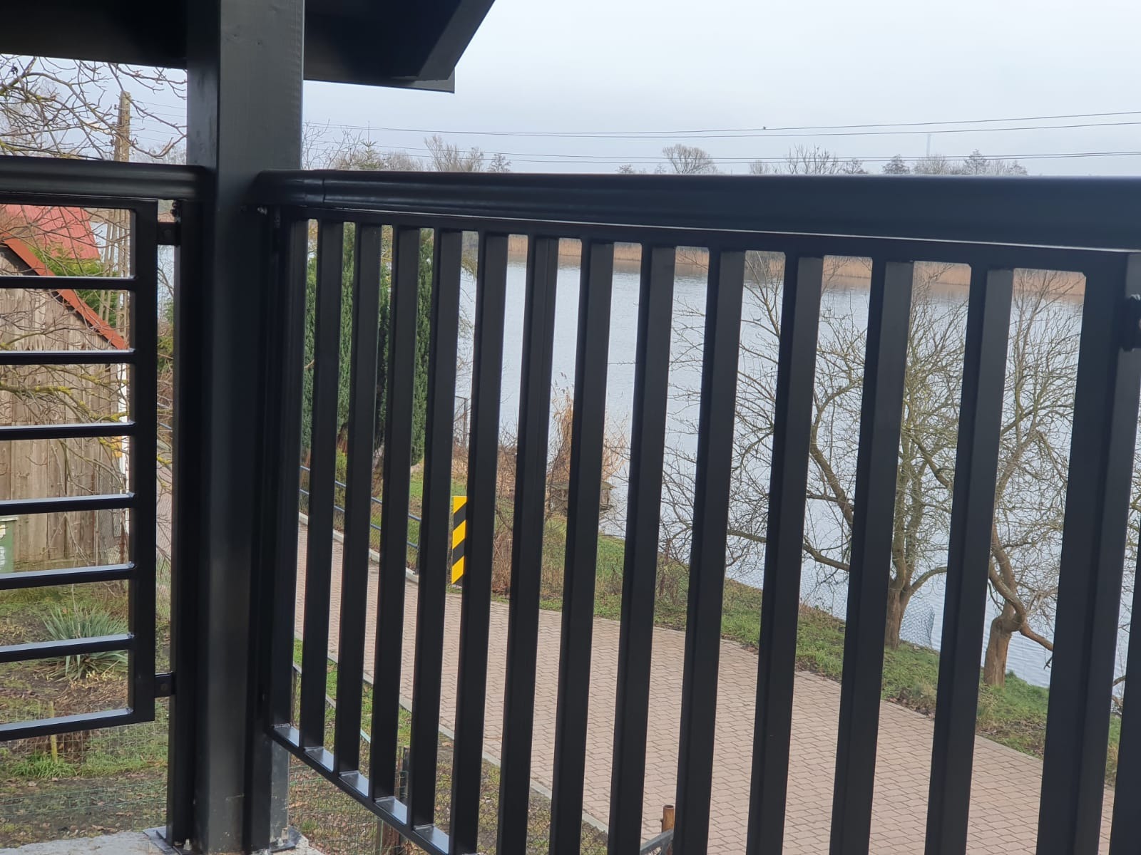 Czarne, pionowe pręty balustrady balkonowej z widokiem na jezioro i ścieżkę spacerową, pochmurne niebo w tle.