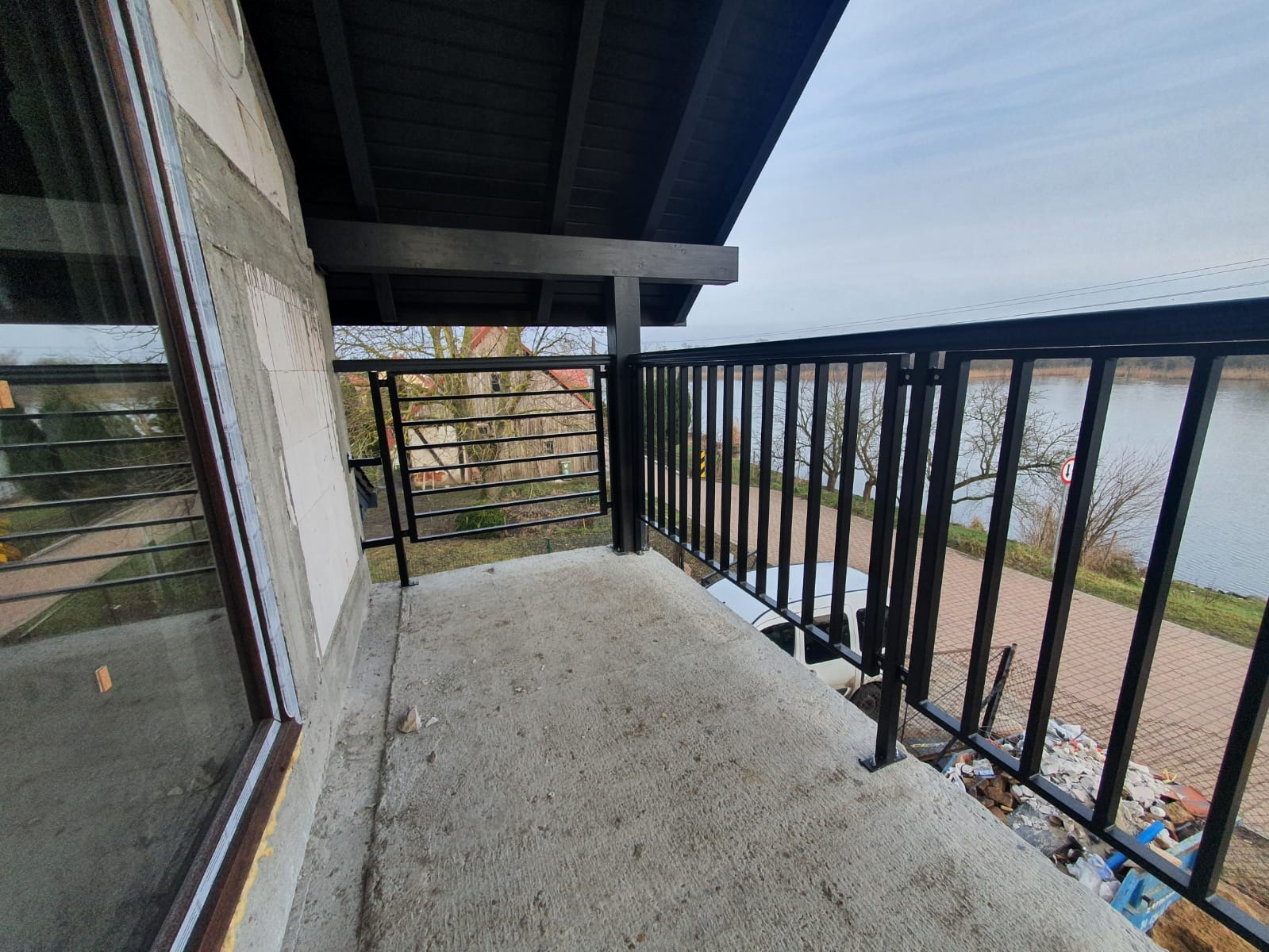 Balkon w trakcie budowy z zamontowaną czarną, metalową balustradą z pionowych szczebli, widok na rzekę i częściowo widoczny samochód na zewnątrz.