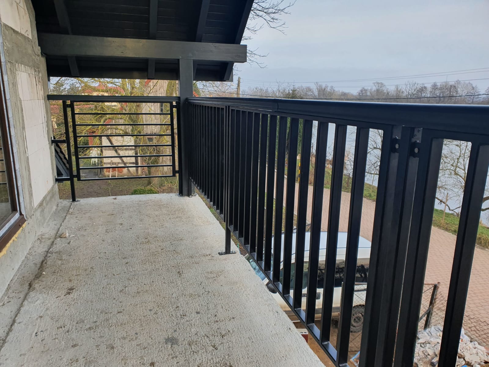 Czarna, stalowa balustrada balkonowa o minimalistycznym designie, widok z zewnątrz na krajobraz z drzewami i zabudowaniami, balkon w stanie surowym.