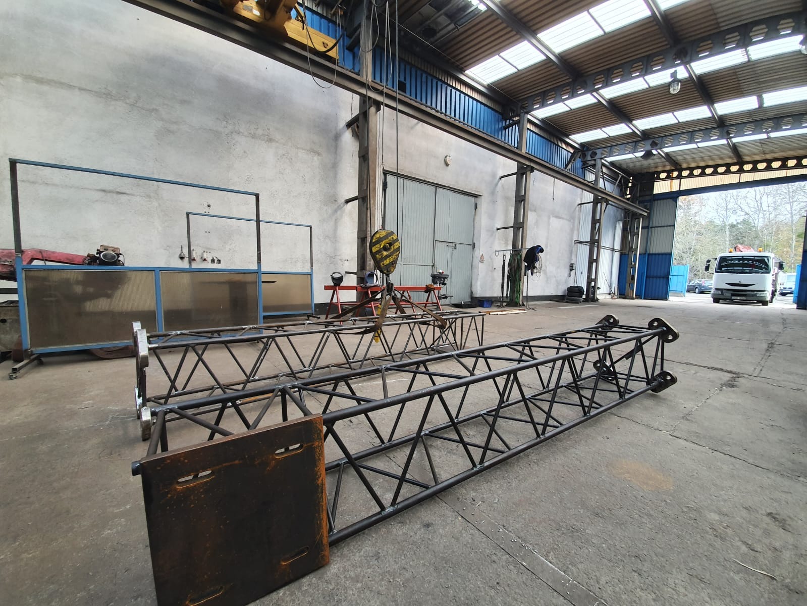 Dwie metalowe konstrukcje kratownicowe, prawdopodobnie do banerów reklamowych, leżące na betonowej podłodze w hali produkcyjnej. Widoczny suwnicowy hak, elementy wyposażenia warsztatu i brama...